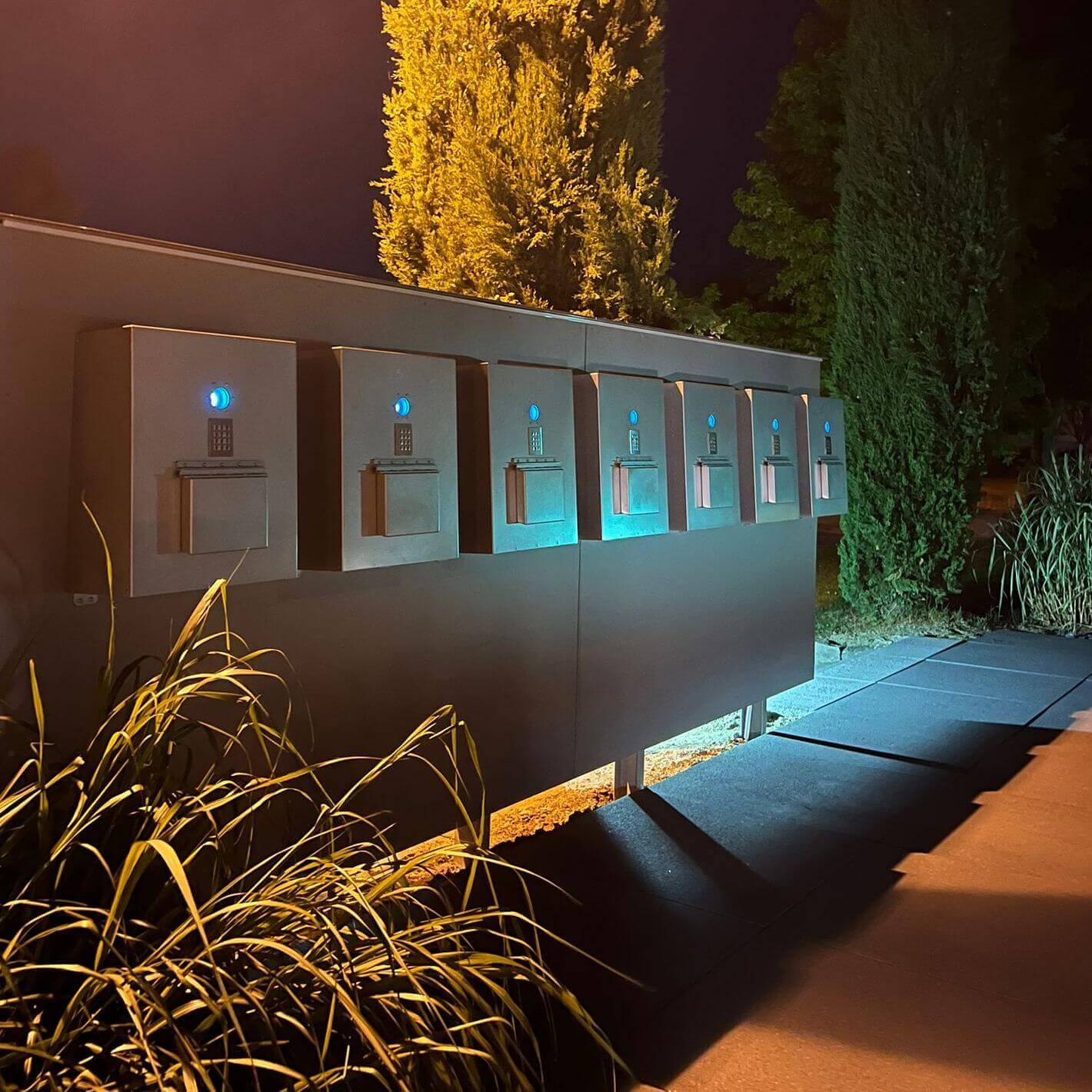 Automatisierte Schlüsselausgabe Livionkey mit Zahlenschloss bei Nacht im Freien Mehrere beleuchtete Schlüsseldepots mit Zahlenschloss in einer Außenanlage, fotografiert am Abend neben hohen Büschen.