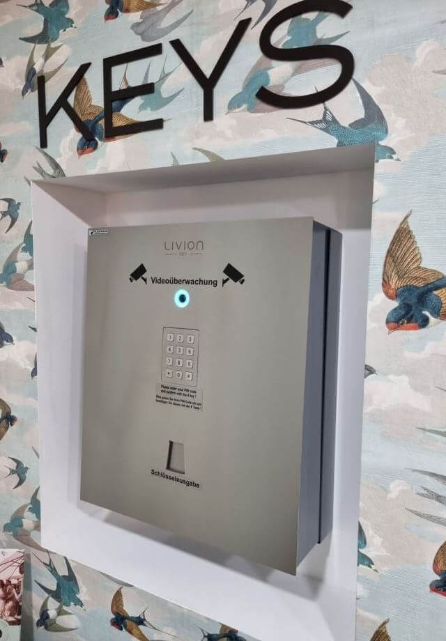 Moderner Schlüsselkasten mit Videoüberwachung und Zahlencode unter großer 'Keys' Beschriftung an einer bunt gemusterten Wand.