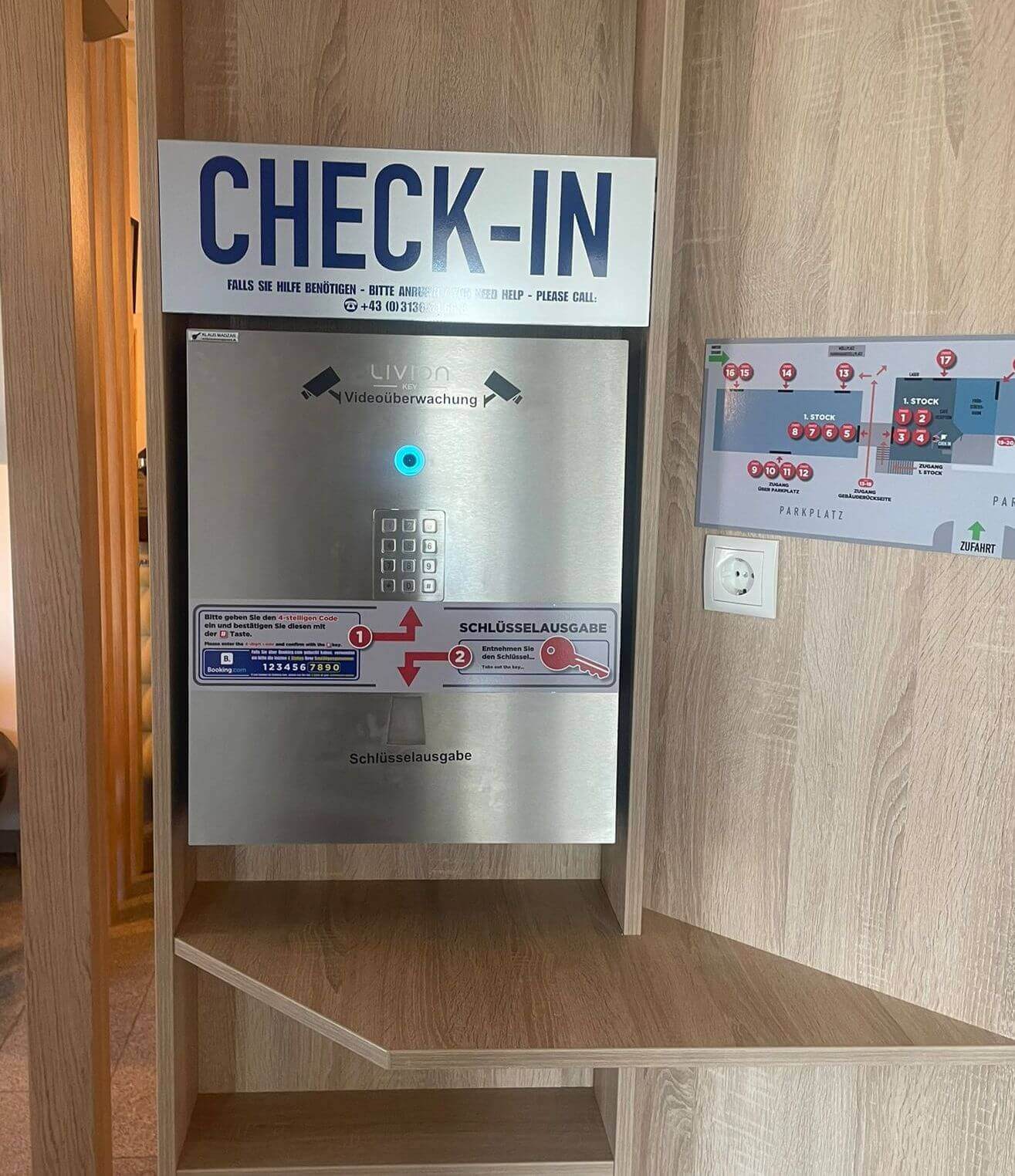 Hotel Check-in Terminal mit Schlüsselausgabe und Videokamera Check-in Automat für Hotels mit Schlüsselausgabe, Videokamera und Bedienungsanleitung, daneben ein Lageplan an der Wand