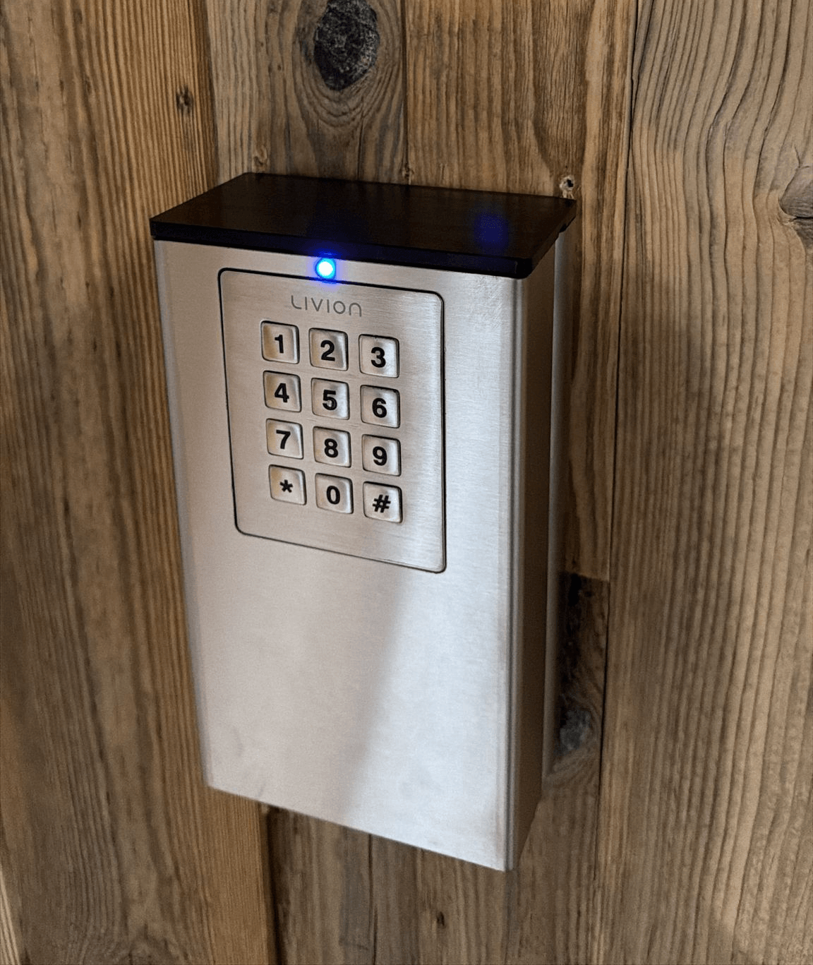 Elektronisches LIVION Zahlencode-Türschloss aus Edelstahl mit beleuchteter Tastatur Wandmontiertes elektronisches LIVION Türschloss aus Edelstahl mit numerischer Code-Tastatur und blauer Status-LED auf Holzwand