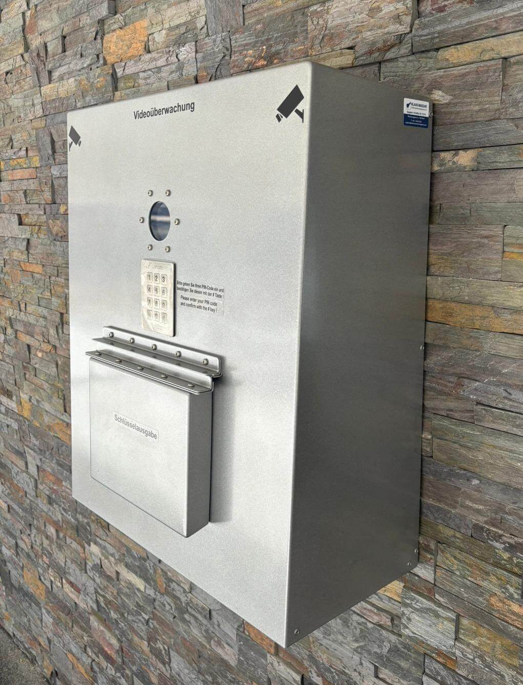 Schlüsselausgabe-Automat aus Edelstahl mit Zahlencode an einer Hauswand, überwacht durch Videokameras Schlüsselausgabe-Automat aus Metall mit Tastenfeld und Kamera unter dem Hinweis ‚Videoüberwachung‘ an einer Steinfassade