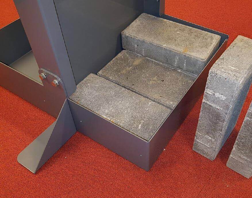 Nahaufnahme eines Stahlrahmens, in dem mehrere graue Betonsteine als Ballast eingesetzt sind, daneben weitere Steine auf rotem Teppich