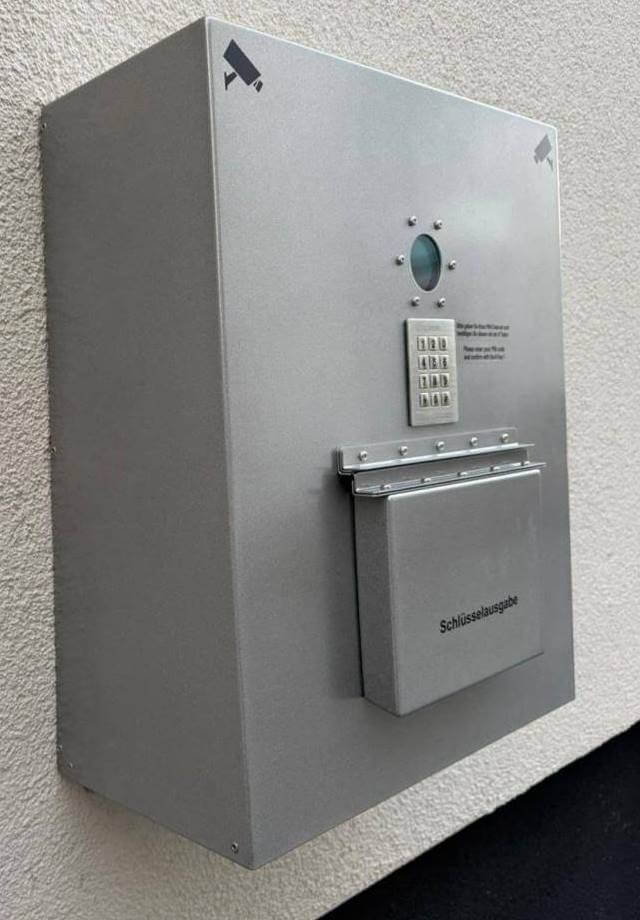 Ein grauer Schlüsselausgabe-Automat an der Wand mit Zahlencode-Tastatur und Überwachungskamera-Symbolen.
