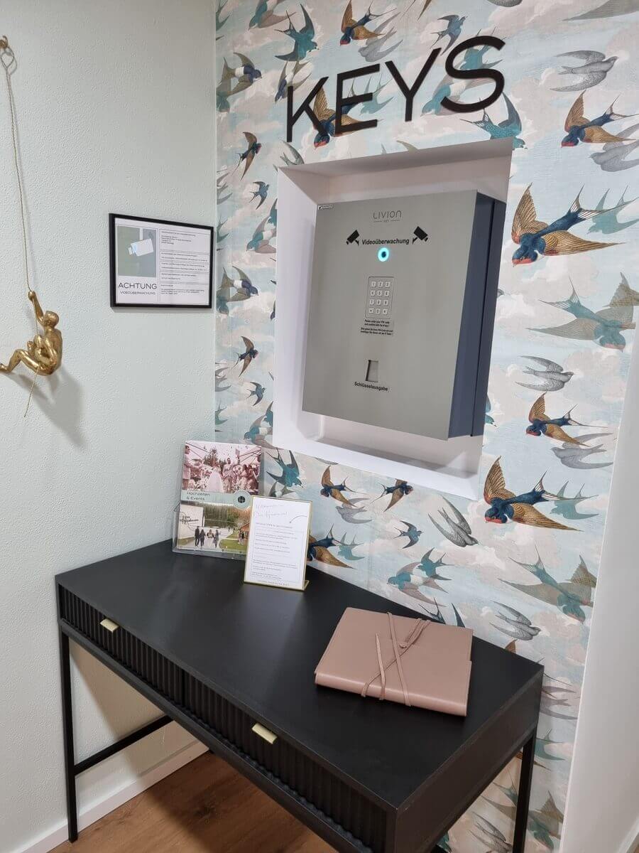 Schlüsselausgabe mit sicherem Schlüsselkasten und Tisch in stilvollem Eingangsbereich Foto eines Eingangsbereichs mit elektronischem Schlüsselkasten, dekorativer Tapete mit Vögeln, schwarzem Tisch, Infomaterial und goldener Kletterfigur an der Wand.