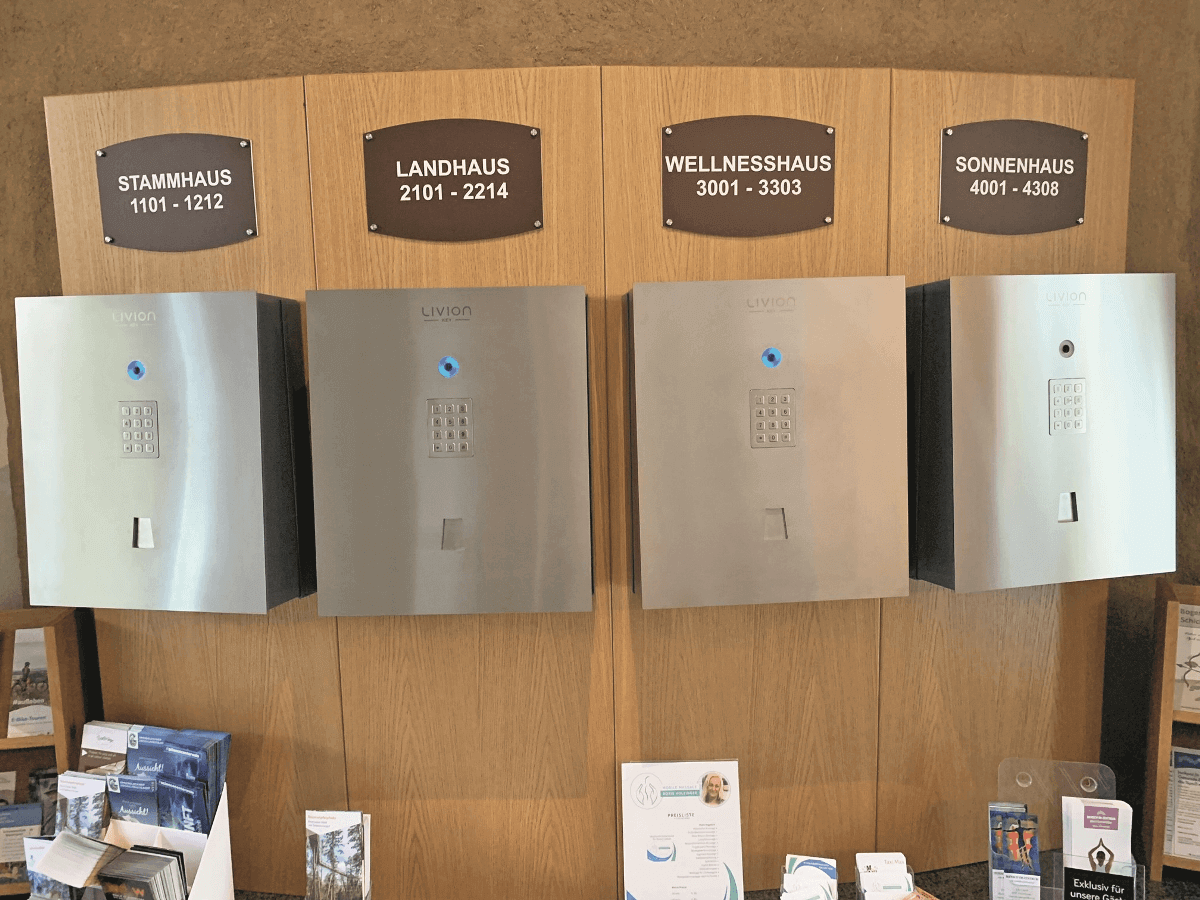 Vier Schlüsselausgabeboxen mit Zahlencodes unter Hausbezeichnungen Stammhaus, Landhaus, Wellnesshaus und Sonnenhaus Schlüsselausgabeboxen mit Tastenfeldern für Stammhaus, Landhaus, Wellnesshaus und Sonnenhaus in einer Unterkunft, darunter Prospekte auf einem Tresen