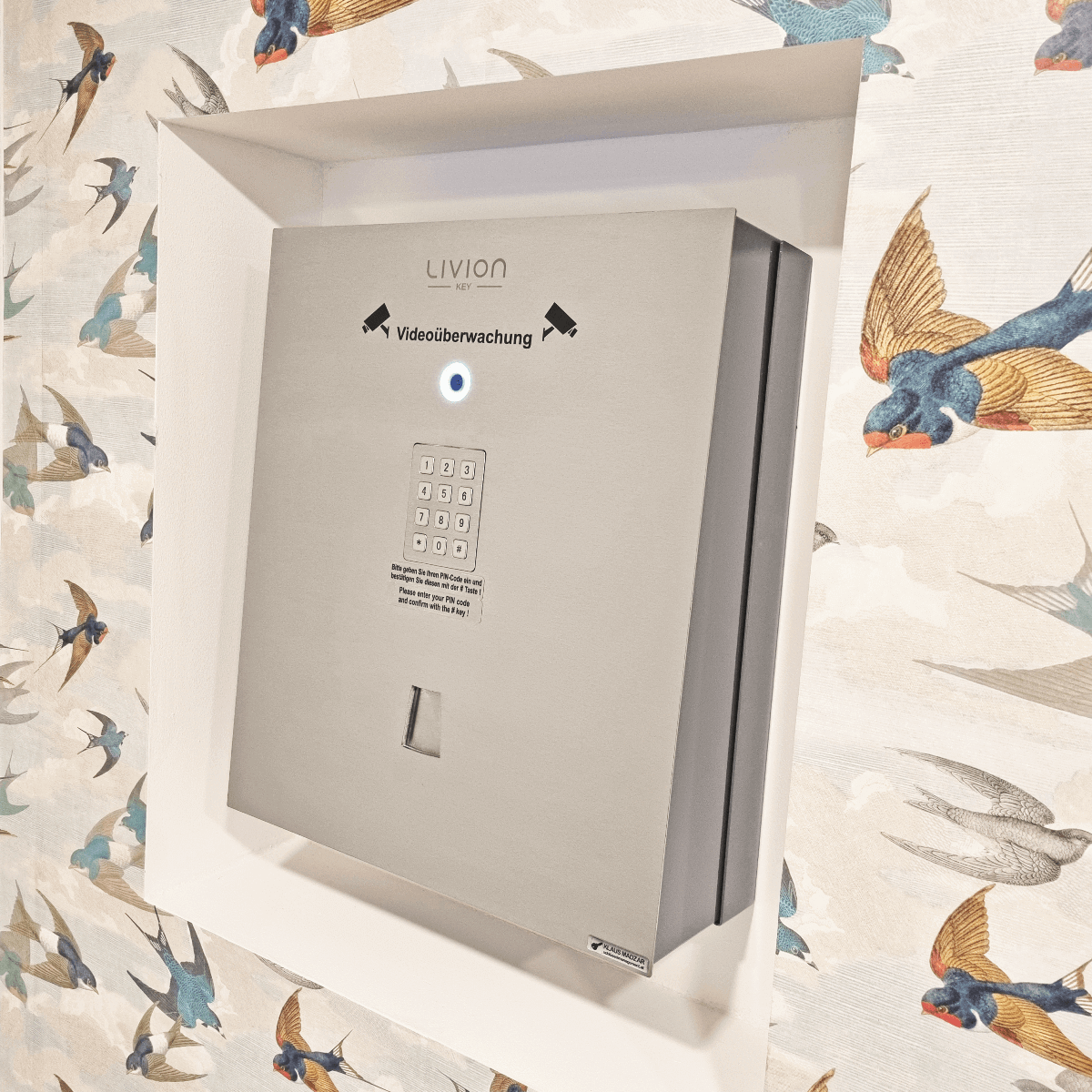 Livion Key Tresor mit Videoberwachung und PIN-Eingabe – Wandmontage, sicherer Aufbewahrungsort Sicherer Livion Key Tresor mit Videoberwachung und PIN-Code-Eingabe an einer Wand mit Vogelmotiv-Tapete montiert