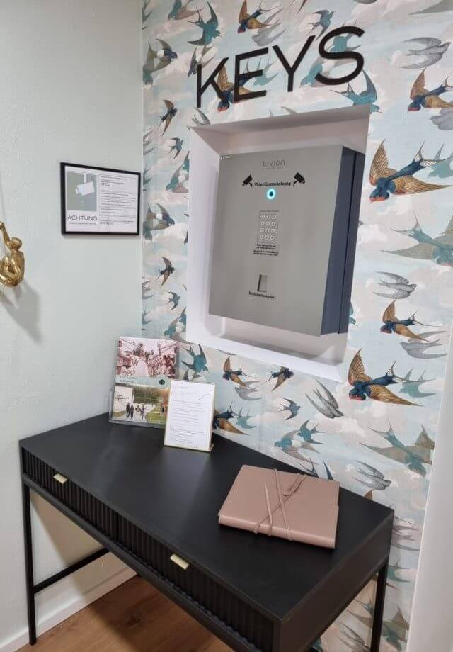 Raumecke mit Schlüsselsafe in die Wand eingelassen, umgeben von Vogel-Tapete, und einem Schreibtisch mit Dekorationen.