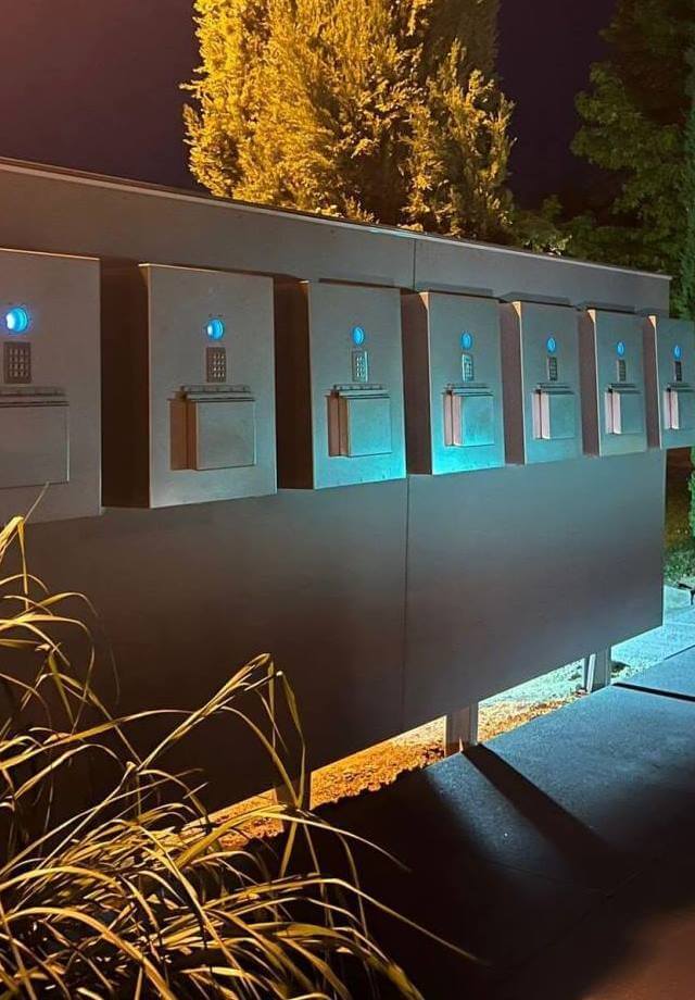 Mehrere Briefkästen in einer beleuchteten Anlage aus Metall bei Nacht, umgeben von Pflanzen