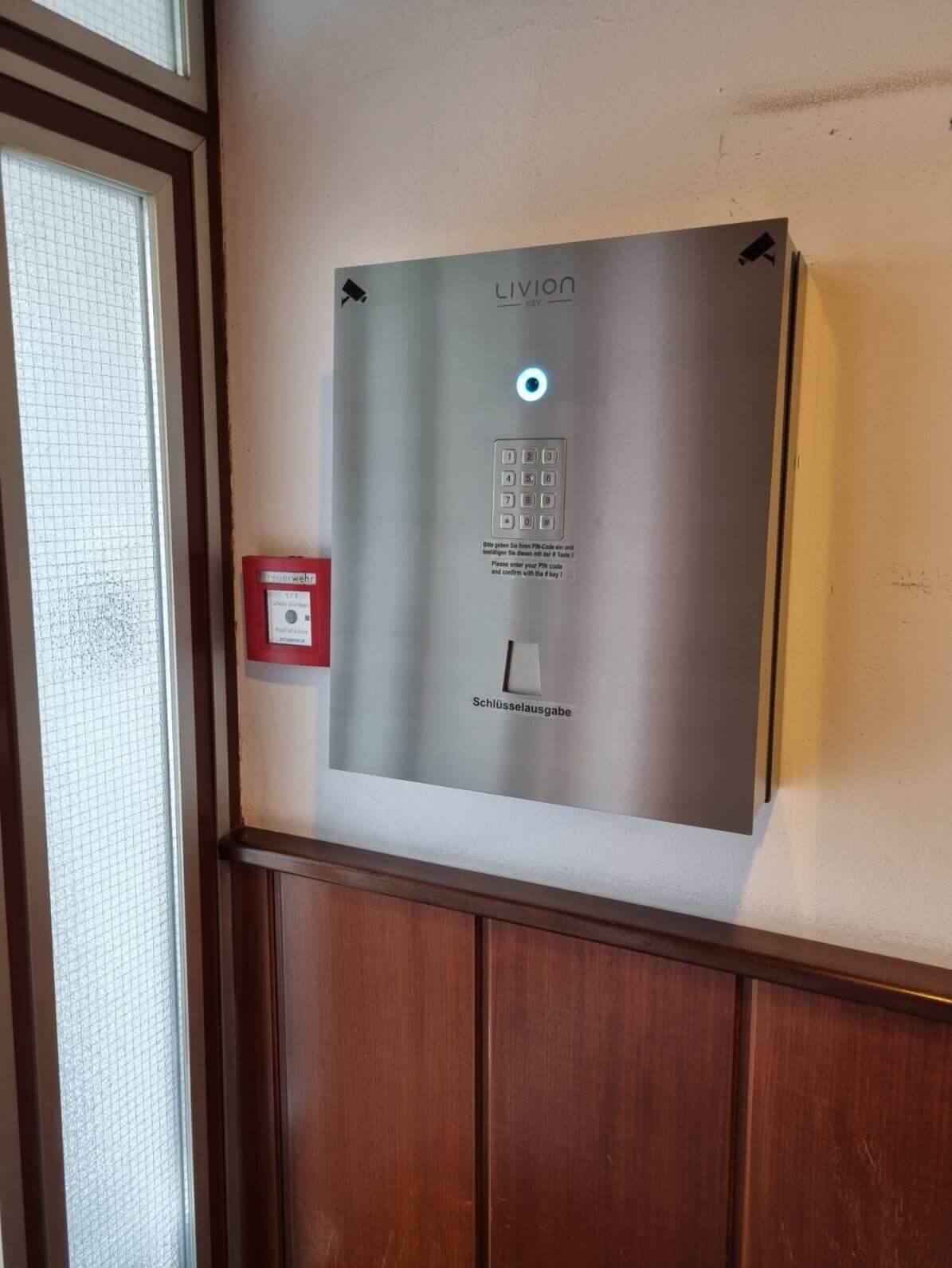 Elektronische Schlüsselausgabe LIVION Key mit PIN-Pad an einer Wand montiert Wandmontierte Schlüsselausgabeanlage LIVION Key aus Edelstahl mit beleuchtetem Tastenfeld und PIN-Eingabemöglichkeit neben einem Feueralarm-Kasten.