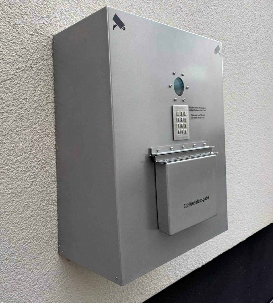 Elektronische Schlüsselausgabebox mit Code-Eingabe und Überwachungskamerasymbolen an einer Wand Wandmontierte, silberne Schlüsselausgabebox mit Tastaturfeld für Zahlencode und Überwachungskamerasymbolen, geeignet für sichere Schlüsselübergabe